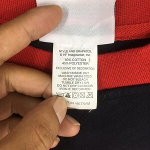 Puede incluir: Primer plano de una etiqueta blanca con texto negro que detalla las instrucciones de cuidado y la composici&oacute;n de la tela. La etiqueta est&aacute; adherida a una prenda roja y negra. La etiqueta dice "STYLE AND GRAPHICS, &copy; VF Imagewear, Inc. 60% ALGOD&Oacute;N 40% POLI&Eacute;STER EXCLUSIVO DECORACI&Oacute;N LAVAR DEL REV&Eacute;S LAVAR A M&Aacute;QUINA EN FR&Iacute;O NO USAR LEJ&Iacute;A SECAR EN SECADORA A BAJA TEMPERATURA NO LIMPIAR EN SECO O PLANCHAR SI EST&Aacute; DECORADO HECHO EN VIETNAM."