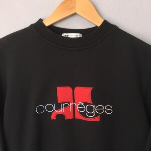 May include: Black sweatshirt with the Courr&egrave;ges logo embroidered in red and white. The logo features a stylized design with the brand name below. The sweatshirt is hanging on a wooden hanger.