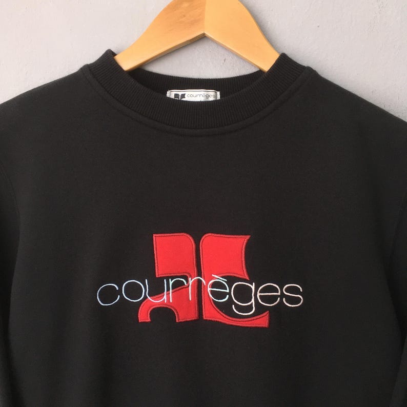 May include: Black sweatshirt with the Courr&egrave;ges logo embroidered in red and white. The logo features a stylized design with the brand name below. The sweatshirt is hanging on a wooden hanger.
