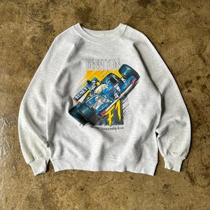 Op de afbeelding: Lichtgrijs sweatshirt met een kleurrijke afbeelding van een Benetton Formule 1-raceauto. Het ontwerp bevat het Benetton-logo en de tekst "World Championship Team". De sweatshirt heeft een ronde hals en lange mouwen.
