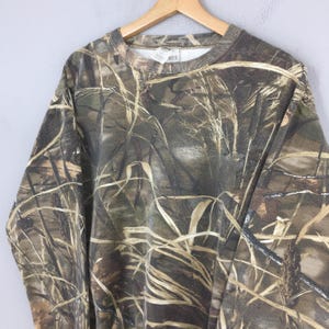 May include: A camouflage-patterned sweatshirt hangs on a wooden hanger. The design features a blend of brown, green, and tan hues, mimicking a natural outdoor environment. The long-sleeved garment has a crew neck and a ribbed collar and cuffs.
