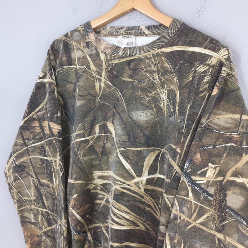 May include: A camouflage-patterned sweatshirt hangs on a wooden hanger. The design features a blend of brown, green, and tan hues, mimicking a natural outdoor environment. The long-sleeved garment has a crew neck and a ribbed collar and cuffs.