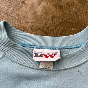 May include: A light blue crewneck sweatshirt featuring a ribbed collar and a white interior tag. The tag displays the text BASSETT-WALKER and BW in red and black lettering, with a secondary tag below indicating size L (42-44) and manufacturing details.
