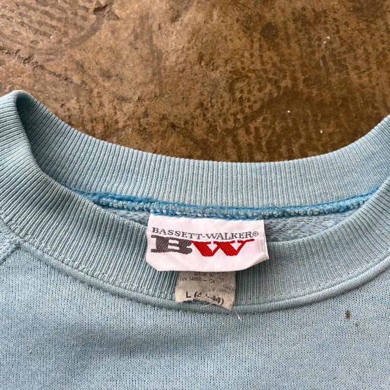 May include: A light blue crewneck sweatshirt featuring a ribbed collar and a white interior tag. The tag displays the text BASSETT-WALKER and BW in red and black lettering, with a secondary tag below indicating size L (42-44) and manufacturing details.