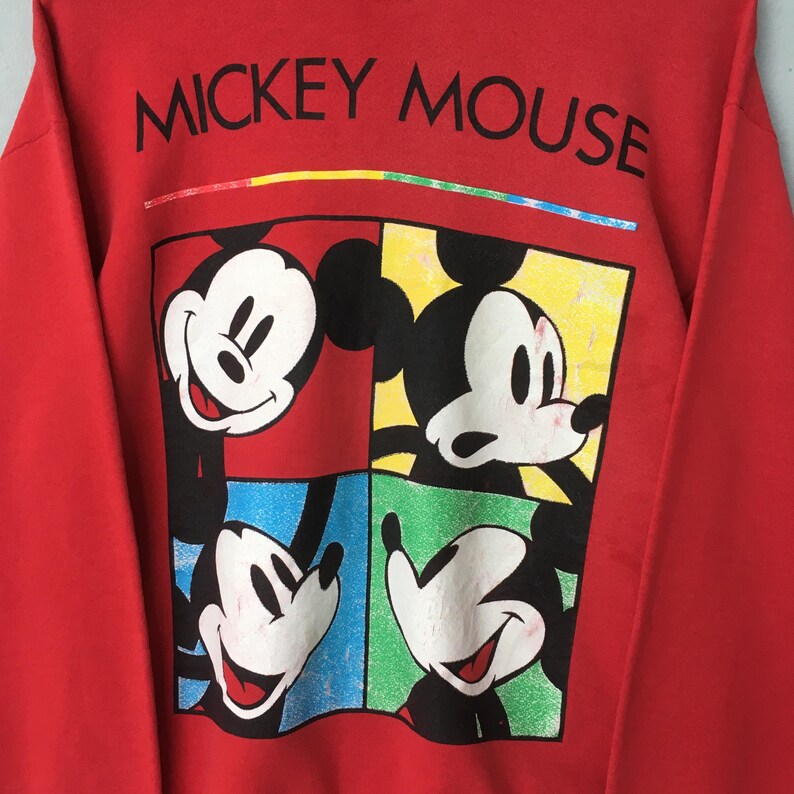 May include: Red sweatshirt featuring a graphic of Mickey Mouse in four different poses, each within a colored square. The words "MICKEY MOUSE" are printed across the top in black. A colorful stripe is above the text.