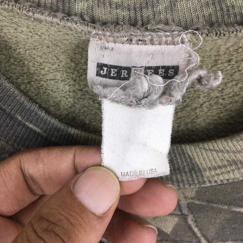 May include: Close-up of a worn, olive green sweatshirt tag. The tag reads "JERZEES" in black letters. Below the brand name, the tag reads "MADE IN USA" in black letters. The sweatshirt has a camouflage pattern.