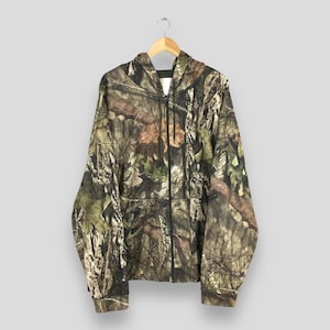 K&ouml;nnte beinhalten: Ein Camouflage-Kapuzenpullover mit Rei&szlig;verschluss und Kapuze, der an einem Holzb&uuml;gel h&auml;ngt. Die Jacke hat ein Muster aus Braun, Gr&uuml;n und Beige, das einer Waldumgebung &auml;hnelt. Der Rei&szlig;verschluss ist schwarz.