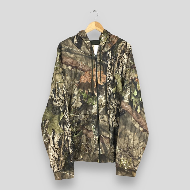 K&ouml;nnte beinhalten: Ein Camouflage-Kapuzenpullover mit Rei&szlig;verschluss und Kapuze, der an einem Holzb&uuml;gel h&auml;ngt. Die Jacke hat ein Muster aus Braun, Gr&uuml;n und Beige, das einer Waldumgebung &auml;hnelt. Der Rei&szlig;verschluss ist schwarz.