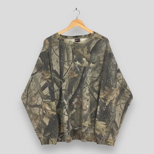 Op de afbeelding: Een sweatshirt met lange mouwen en ronde hals voorzien van een all-over camouflageprint met een realistisch bospatroon van boomschors, takken en gevallen bladeren in bruine, beige en donkergroene tinten. Het kledingstuk hangt aan een houten hanger.