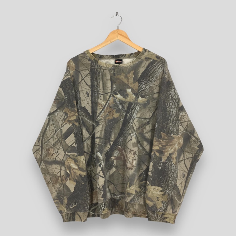 Op de afbeelding: Een sweatshirt met lange mouwen en ronde hals voorzien van een all-over camouflageprint met een realistisch bospatroon van boomschors, takken en gevallen bladeren in bruine, beige en donkergroene tinten. Het kledingstuk hangt aan een houten hanger.