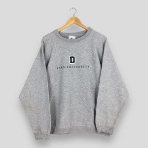 Może przedstawiać: Szara bluza z napisem "DUKE UNIVERSITY" w kolorze granatowym. Nad napisem znajduje się granatowa litera "D". Bluza wisi na drewnianym wieszaku.