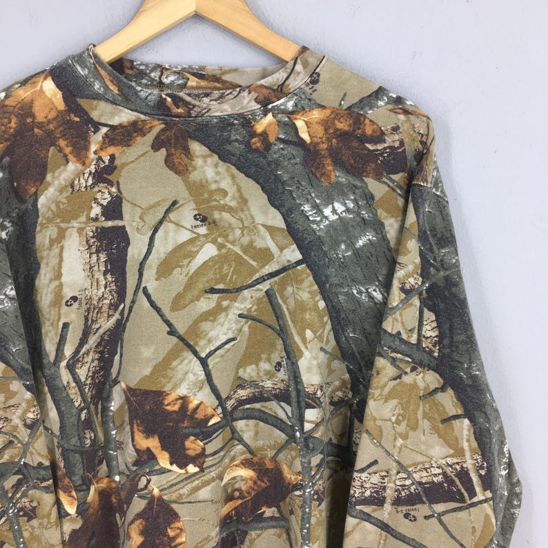 May include: Long-sleeved camouflage shirt in shades of brown, green, and tan. The shirt features a crew neck and a pattern of leaves and branches, typical of hunting apparel.