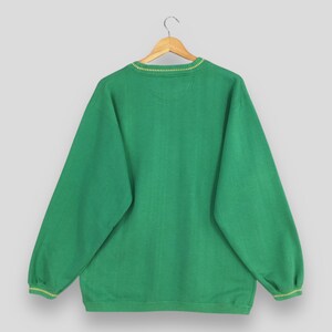 May include: A green sweatshirt with a crew neck and long sleeves, hanging on a wooden hanger. The neckline and cuffs feature a thin yellow trim. The fabric has a subtle textured pattern.