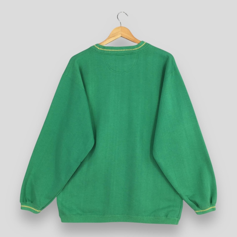 May include: A green sweatshirt with a crew neck and long sleeves, hanging on a wooden hanger. The neckline and cuffs feature a thin yellow trim. The fabric has a subtle textured pattern.