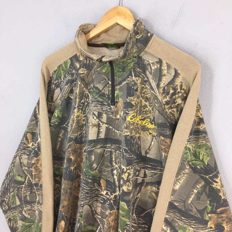 May include: A camouflage-patterned pullover with a quarter-zip closure. The long-sleeved top features a brown collar and shoulder panels, with the brand name "Cabela's" in yellow script. The garment is hanging on a wooden hanger.