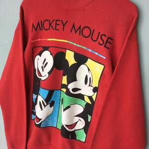 May include: Red sweatshirt featuring a graphic of Mickey Mouse in four different poses. The words "MICKEY MOUSE" are printed across the top in black. A colorful stripe is above the graphic. The sweatshirt is a vintage style.