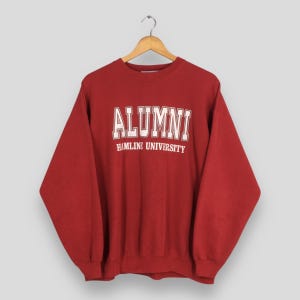 Może przedstawiać: Czerwona bluza z napisem "ALUMNI" w białych literach nad napisem "HAMLINE UNIVERSITY". Bluza wisi na drewnianym wieszaku na neutralnym tle. Top z długim rękawem to klasyczny styl.