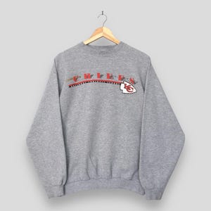 May include: A heather gray crewneck sweatshirt featuring the word CHIEFS in red and black lettering across the chest. Below the text is a black line with the words NATIONAL FOOTBALL LEAGUE and a white arrowhead logo with red KC letters.
