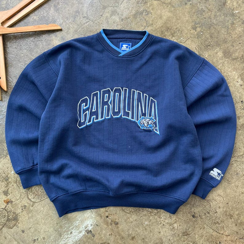 May include: A navy blue long-sleeve sweatshirt featuring the word CAROLINA in white and light blue lettering across the chest. The garment includes a small embroidered mascot logo, a ribbed V-neck collar, and a Starter brand logo on the left sleeve.