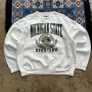 Puede incluir: Sudadera crema con cuello redondo y la inscripción "MICHIGAN STATE" en verde. Debajo, un emblema circular dice "MICHIGAN STATE UNIVERSITY" con "SPARTANS" debajo. Un estilo clásico, ideal para uso informal.