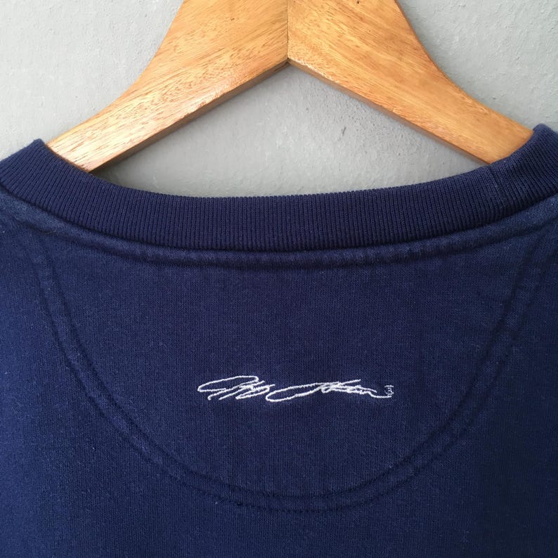 May include: Close-up of a navy blue sweatshirt collar with a ribbed neckline. The back of the sweatshirt features a white embroidered signature. The sweatshirt is hanging on a wooden hanger.