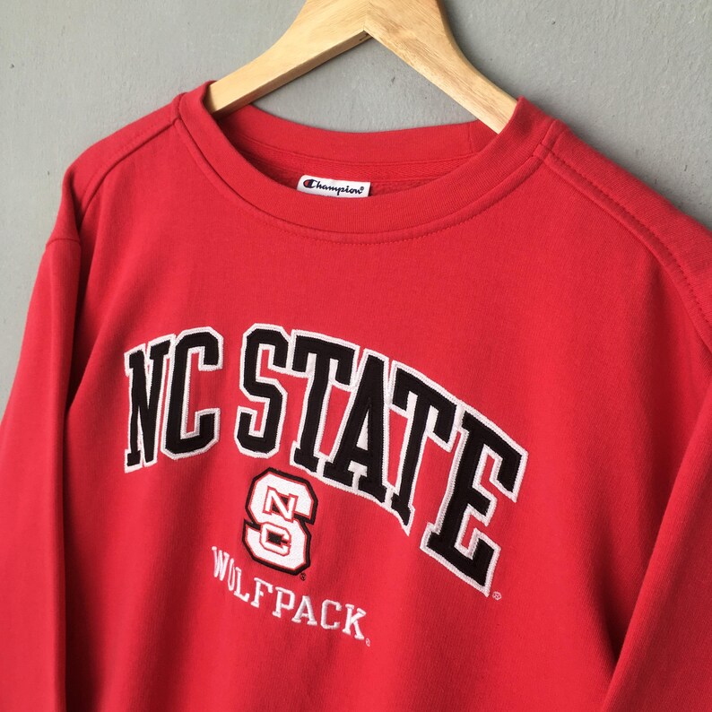 May include: Red Champion sweatshirt with the text "NC STATE" in black and white arched lettering. Below the text is a logo with the letter "S" and the word "WOLFPACK". The sweatshirt is hanging on a wooden hanger.
