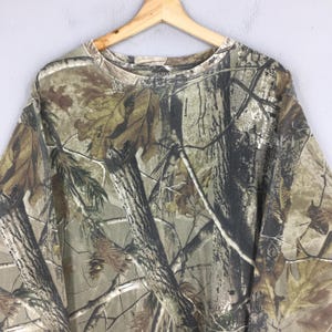 May include: A long-sleeved camouflage shirt with a crew neck, hanging on a wooden hanger. The shirt features a realistic forest pattern in shades of brown, green, and tan, with tree and leaf details. The shirt is made of a soft material.