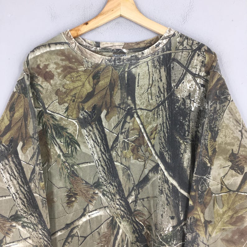May include: A long-sleeved camouflage shirt with a crew neck, hanging on a wooden hanger. The shirt features a realistic forest pattern in shades of brown, green, and tan, with tree and leaf details. The shirt is made of a soft material.