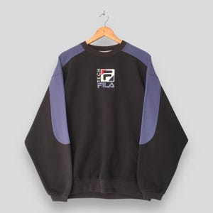 May include: Black sweatshirt with purple shoulder panels and the FILA logo. The logo is white, red, and blue. The word "TECH" is printed vertically on the left side of the logo. The sweatshirt is hanging on a wooden hanger.