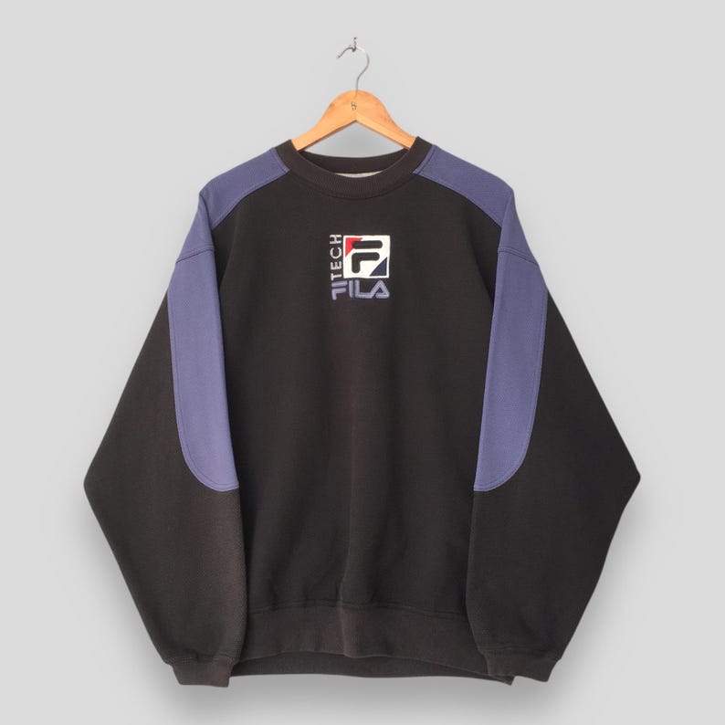 May include: Black sweatshirt with purple shoulder panels and the FILA logo. The logo is white, red, and blue. The word "TECH" is printed vertically on the left side of the logo. The sweatshirt is hanging on a wooden hanger.