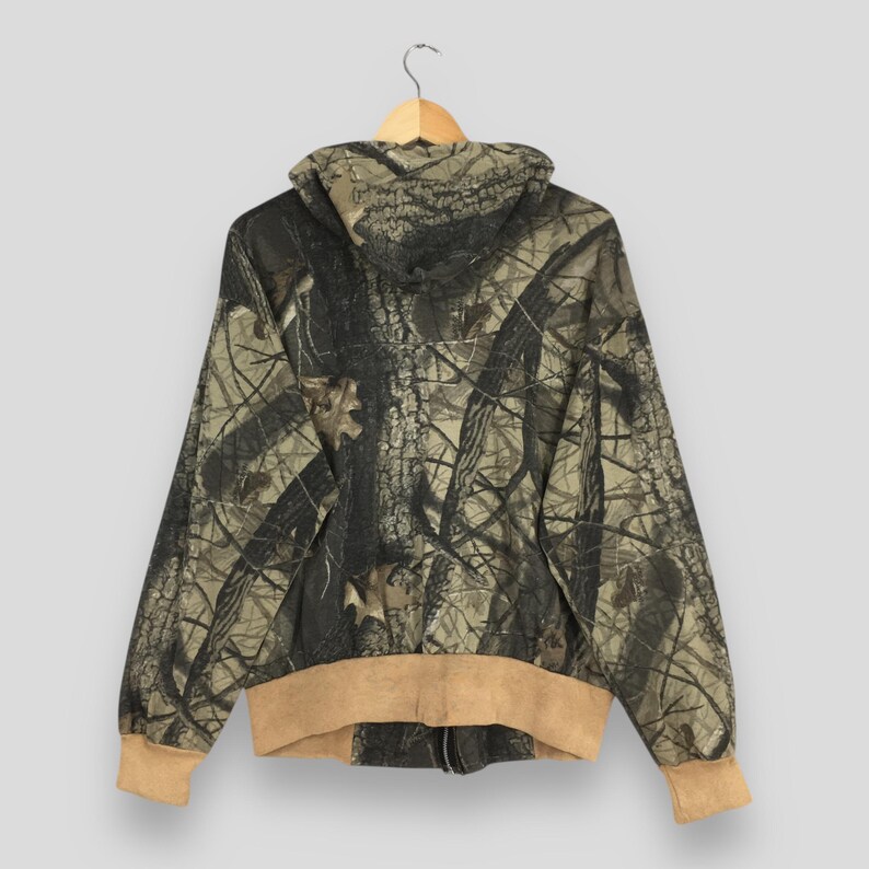 May include: A hooded camouflage jacket with a full zipper, displayed from the back. The jacket features a brown and tan color scheme with a forest pattern. The cuffs and hem are a solid tan color.