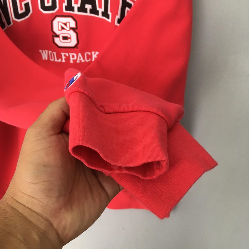 May include: A red Champion sweatshirt with the text "NC STATE" in white arched across the top, and "WOLFPACK" below. The number "20" is in the center. The sweatshirt's cuff is visible.