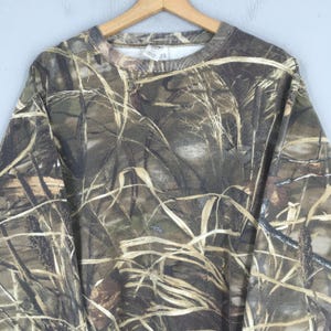 May include: A camouflage-patterned sweatshirt in shades of brown, green, and tan, hanging on a wooden hanger. The design mimics a natural outdoor environment, with a crew neck and long sleeves.