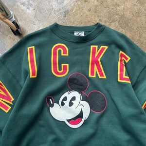 K&ouml;nnte beinhalten: Ein dunkelgr&uuml;nes Sweatshirt mit dem Wort "MICKEY" in roten Buchstaben mit gelben Umrissen. Das Sweatshirt hat eine Grafik von Mickey Mouses Gesicht. Die &Auml;rmel tragen ebenfalls das Wort "MICKEY".