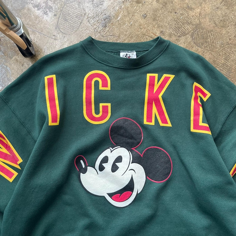 K&ouml;nnte beinhalten: Ein dunkelgr&uuml;nes Sweatshirt mit dem Wort "MICKEY" in roten Buchstaben mit gelben Umrissen. Das Sweatshirt hat eine Grafik von Mickey Mouses Gesicht. Die &Auml;rmel tragen ebenfalls das Wort "MICKEY".
