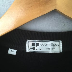 May include: Close-up of a black crew neck shirt with a white label. The label reads "courr&egrave;ges and co" in a sans-serif font. A white "M" size tag is visible. The shirt is hanging on a wooden hanger.