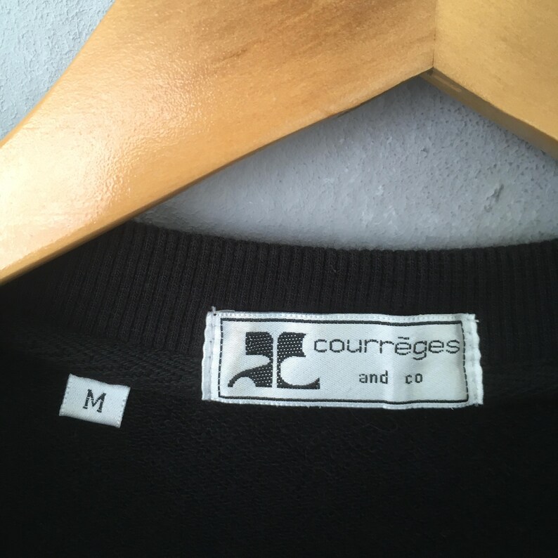 May include: Close-up of a black crew neck shirt with a white label. The label reads "courr&egrave;ges and co" in a sans-serif font. A white "M" size tag is visible. The shirt is hanging on a wooden hanger.