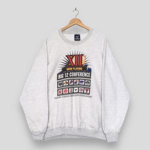 May include: A light gray heathered crewneck sweatshirt featuring a graphic print that reads XII NOW PLAYING OUR FEATURE PRESENTATION BIG 12 CONFERENCE. The design includes a film strip motif with various university logos and small text below.