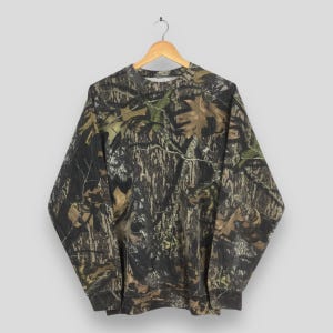 May include: A long-sleeved crewneck sweatshirt featuring a realistic forest camouflage pattern in shades of dark brown, black, tan, and muted green. The garment has a ribbed collar, cuffs, and hem, displaying a detailed print of tree bark and leaves.