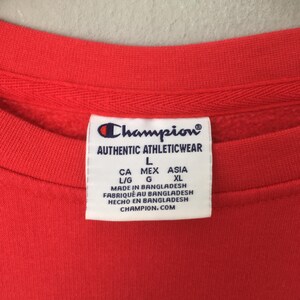 May include: Close-up of a red Champion sweatshirt. The white tag displays the Champion logo and text: AUTHENTIC ATHLETICWEAR, L, CA MEX ASIA, L/G G, XL, MADE IN BANGLADESH, FABRIQU&Eacute; AU BANGLADESH, HECHO EN BANGLADESH, CHAMPION.COM.