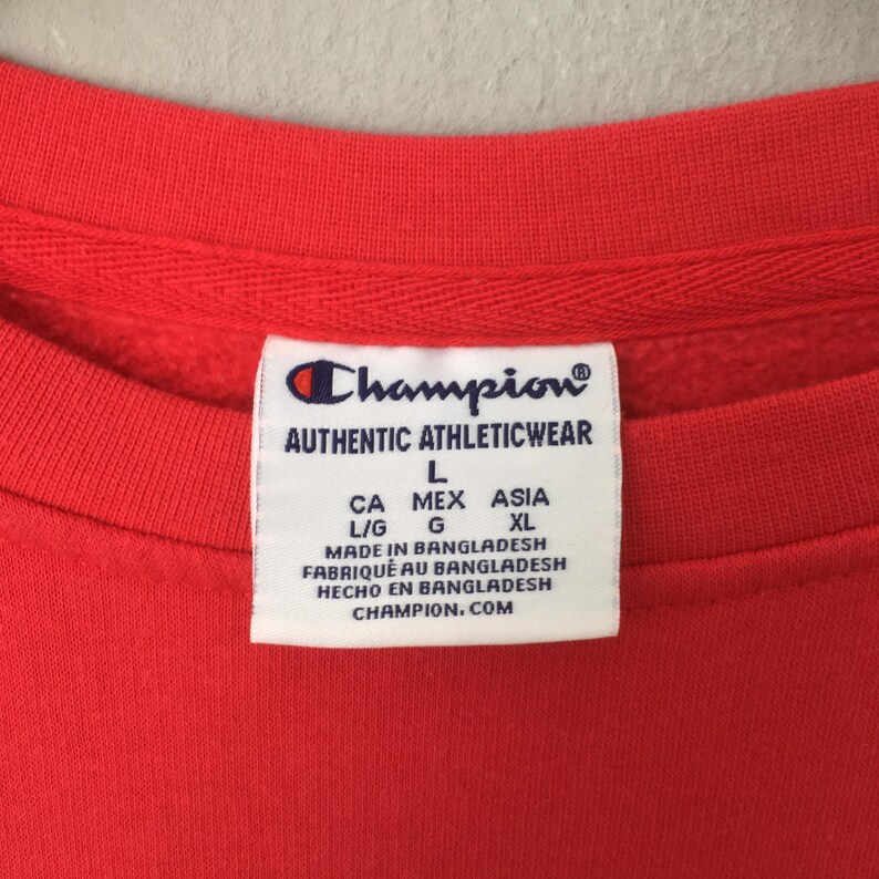 May include: Close-up of a red Champion sweatshirt. The white tag displays the Champion logo and text: AUTHENTIC ATHLETICWEAR, L, CA MEX ASIA, L/G G, XL, MADE IN BANGLADESH, FABRIQU&Eacute; AU BANGLADESH, HECHO EN BANGLADESH, CHAMPION.COM.