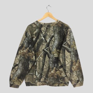 Puede incluir: Una sudadera con estampado de camuflaje cuelga de una percha de madera. La parte superior de manga larga presenta un estampado forestal realista en tonos marrones, verdes y beiges. La sudadera de cuello redondo est&aacute; hecha de un material suave.
