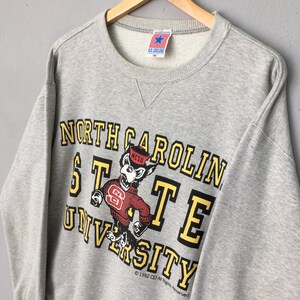 Puede incluir: Una sudadera gris jaspeado de cuello redondo con el texto North Carolina State University en letras amarillas y negras. Un gr&aacute;fico de una mascota de lobo con un su&eacute;ter rojo con el logo NC est&aacute; centrado en el frente. Una etiqueta U.S. College est&aacute; en el cuello.