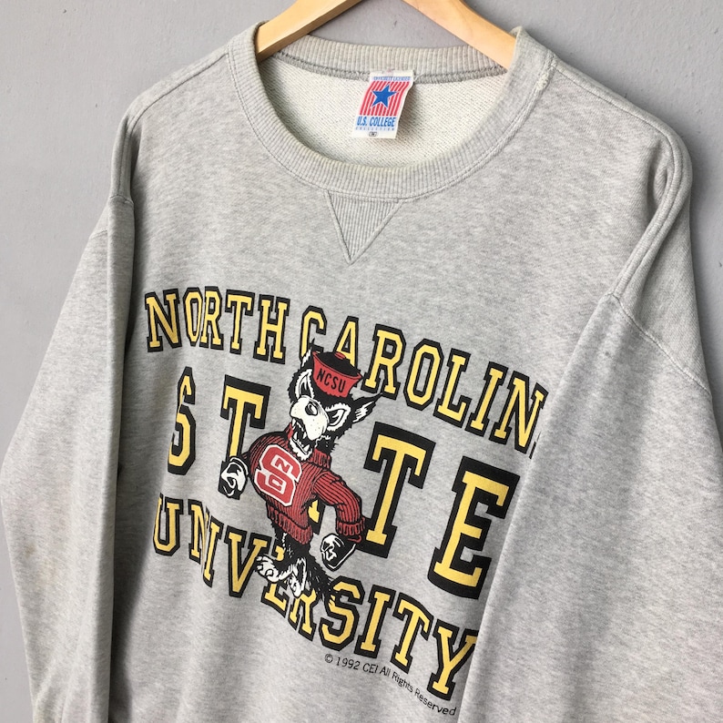 Puede incluir: Una sudadera gris jaspeado de cuello redondo con el texto North Carolina State University en letras amarillas y negras. Un gr&aacute;fico de una mascota de lobo con un su&eacute;ter rojo con el logo NC est&aacute; centrado en el frente. Una etiqueta U.S. College est&aacute; en el cuello.