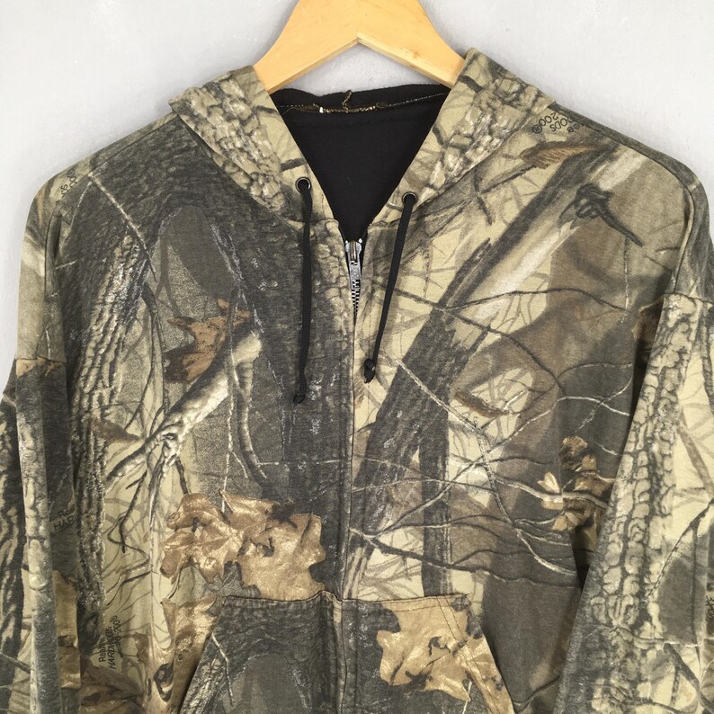 May include: A camouflage-patterned zip-up hoodie with a drawstring hood and a black interior. The jacket features a front pocket and a full-length zipper. The color palette includes shades of brown, green, and tan, creating a realistic woodland camouflage design.