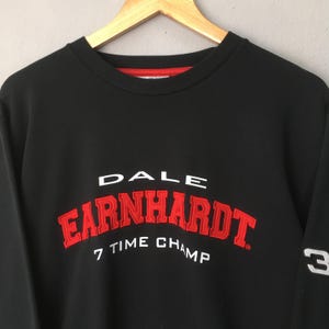 May include: Black sweatshirt with the name "EARNHARDT" in red, arched lettering. The text "DALE" is above, and "7 TIME CHAMP" is below. The number "3" is on the sleeve. The sweatshirt has a red neckline.