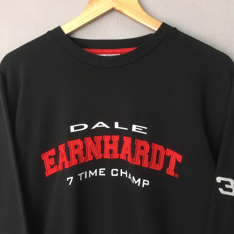 May include: Black sweatshirt with the name "EARNHARDT" in red, arched lettering. The text "DALE" is above, and "7 TIME CHAMP" is below. The number "3" is on the sleeve. The sweatshirt has a red neckline.
