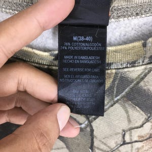 Puede incluir: Primer plano de una etiqueta de ropa en una sudadera con estampado de camuflaje. La etiqueta indica la talla M (38-40) y la composici&oacute;n de la tela: 76% algod&oacute;n, 24% poli&eacute;ster. Tambi&eacute;n indica "Made in Bangladesh" e instrucciones de cuidado.