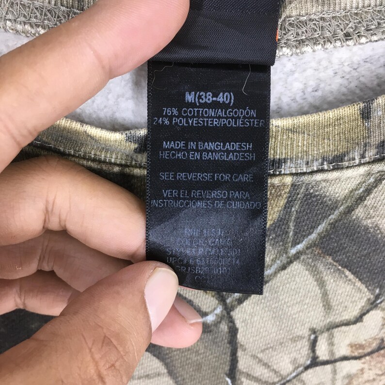 Puede incluir: Primer plano de una etiqueta de ropa en una sudadera con estampado de camuflaje. La etiqueta indica la talla M (38-40) y la composici&oacute;n de la tela: 76% algod&oacute;n, 24% poli&eacute;ster. Tambi&eacute;n indica "Made in Bangladesh" e instrucciones de cuidado.