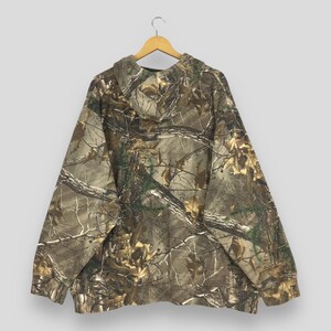 Op de afbeelding: Een camouflage hoodie met een bospatroon in bruin, groen en beige. De hoodie hangt aan een houten hanger tegen een lichtgrijze achtergrond. Het heeft een capuchon met trekkoord en een kangoeroezak.
